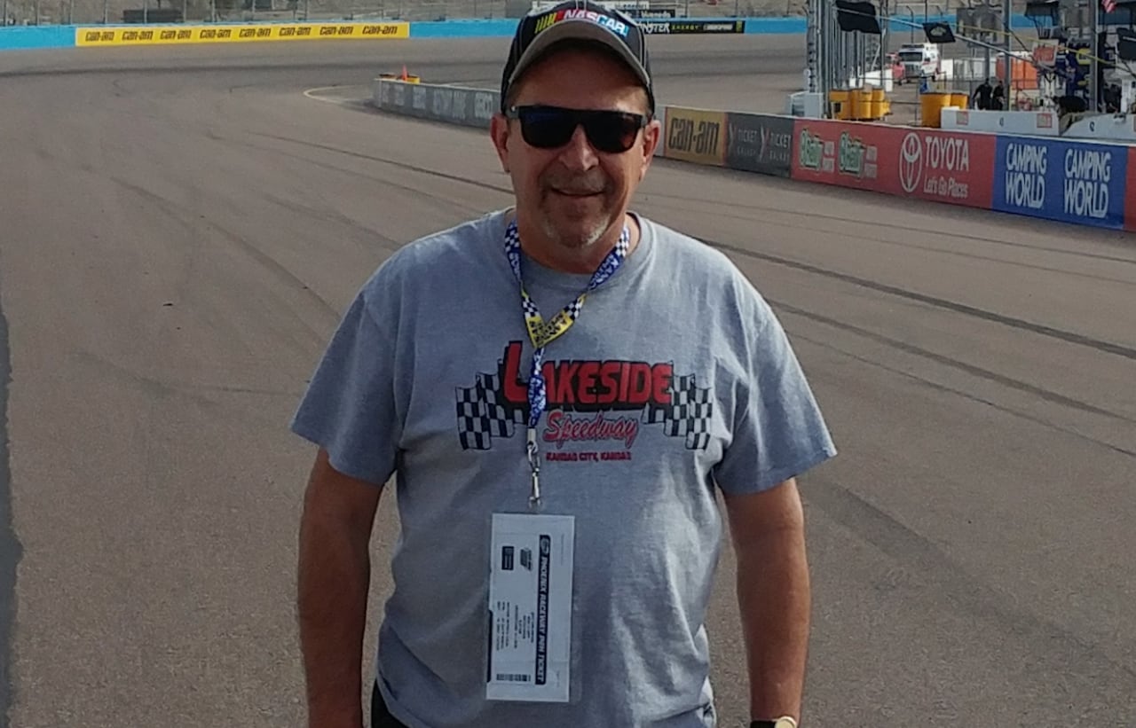 A man in a grey t-shirt and baseball cap stands in the middle of a race track speedway.