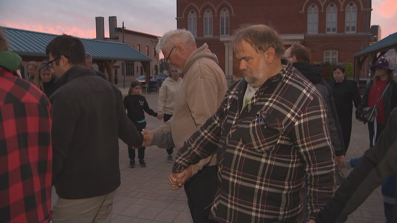 Adults, kids and seniors are shown holding hands and standing in a circle outside Belleville City hall as the sun sets.
