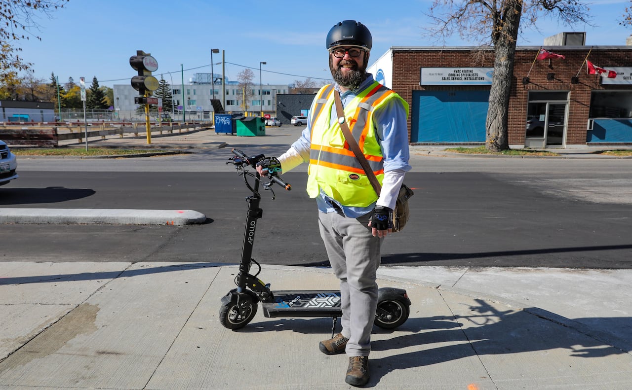 A man with a beard wears a bright yellow and orange vest and a helmet while standing next to an electric scooter beside a city street