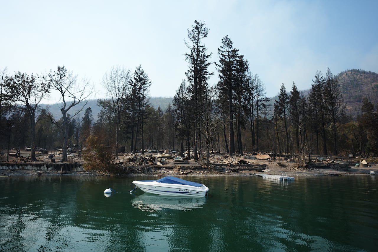 A white speedboat floats in a lake in front of properties that have burned down.
