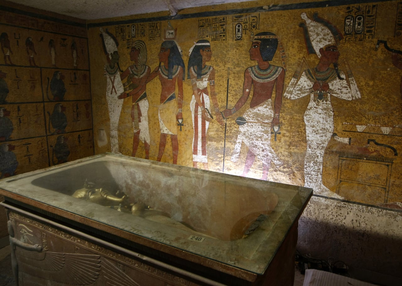 A golden sarcophagus is shown lying in an elaborate burial chamber. The wall behind it is colourfully painted with figures.