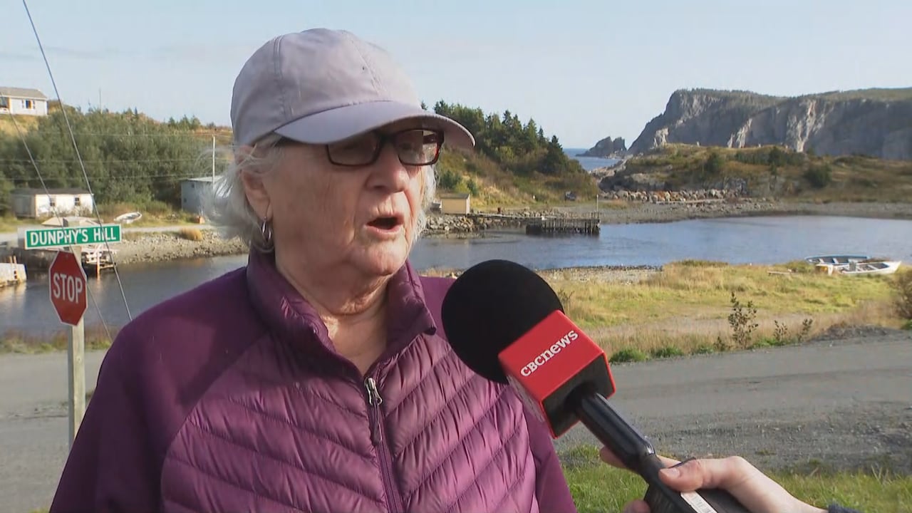 A woman wearing a baseball cap stands on a single-lane roadside, speaking into a CBC microphone.