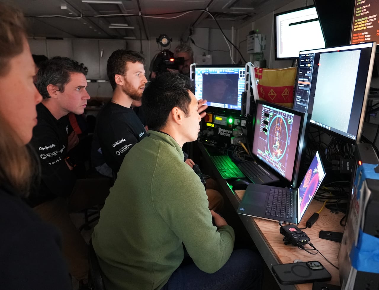 Four people gather around several computer screens as another man with a video camera records.