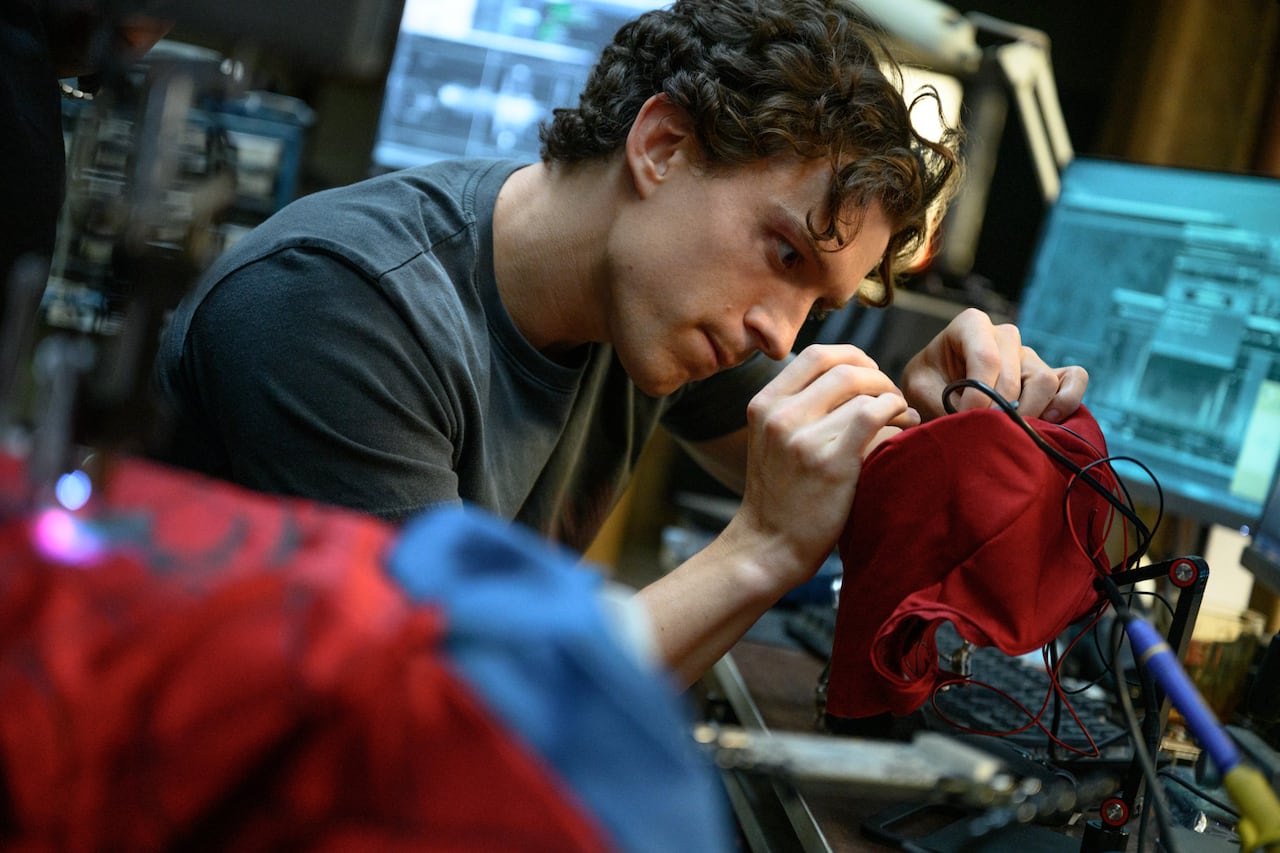 Tom Holland as Peter Parker works on his Spider-Man costume, we see mask and computers on a work bench.
