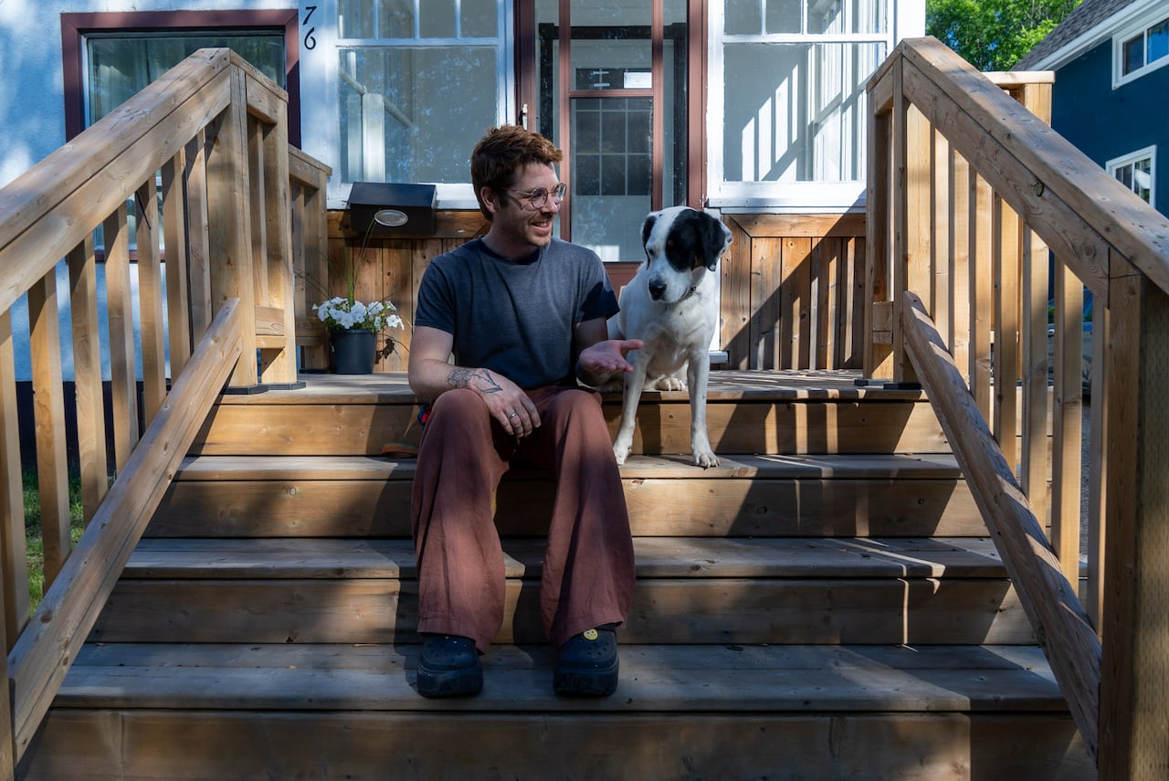 A man with glasses, short red hair and scruffy facial hair sits on the wooden steps of a house next to a black and white dog.