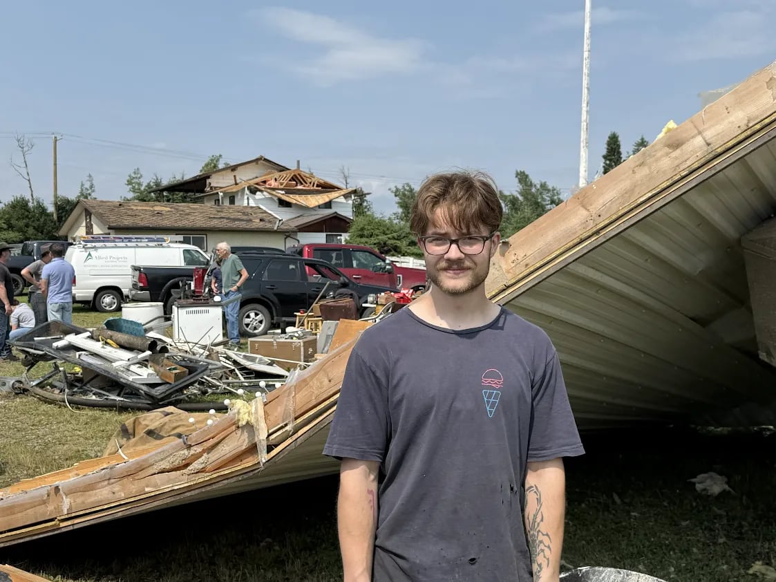 A man with brown hair and a bard, wearing glasses and a t-shirt, stands in front of a home damaged by a tornado.