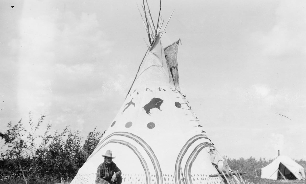 A black and white photo of a teepee with a painted cover. A man in a Western hat crouches on one knee in front of it.