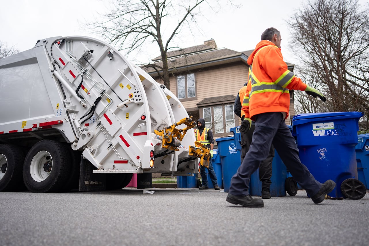 City workers in orange vests work with recycling bins on the road beside a white dump truck