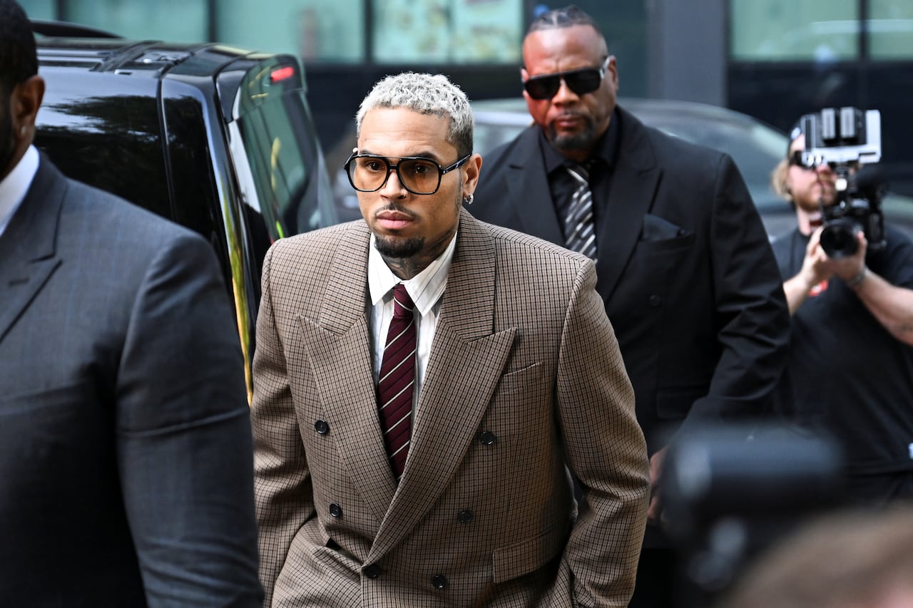 A man in a brown suit and large sunglasses is shown walking near a number of men in black suits and black sunglasses.