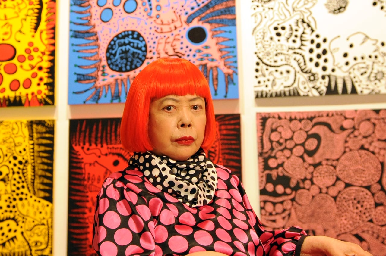 A picture of Yayoi Kusama wearing a polka dot dress in front of her paintings that has swirls, polka dots, etc.