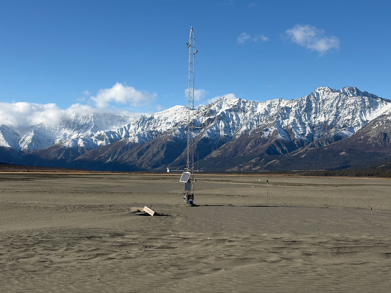 A tall monitoring tower with a solar panel stands on the sandy Ä'äy Chù Valley floor, with snow-covered mountains behind it.