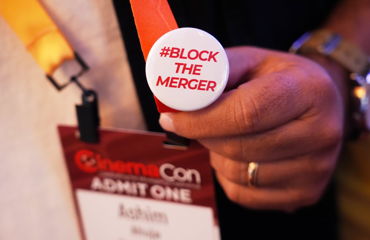 A close-up image of a hand holding a button with the phrase "#BLOCK THE MERGER," pinned to a lanyard from the CinemaCon film industry conference.