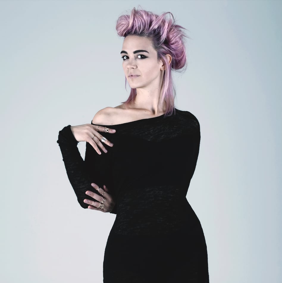 A woman with pink hair in a black dress holding her arms to one side.