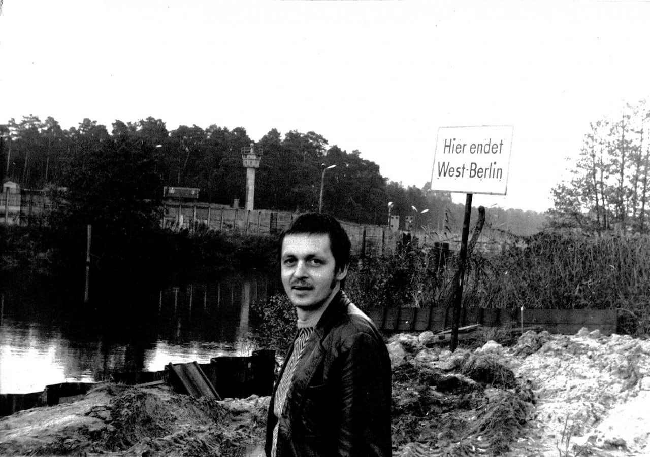 A man stands in front of the Spree river in black and white, with the Berlin wall behind him.