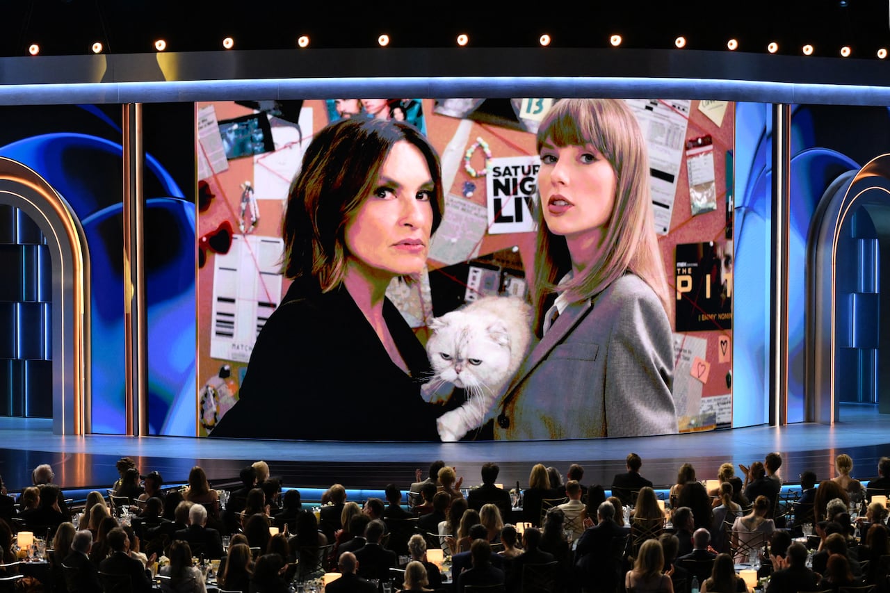 A screen shows two women in suits in front of a crowd.