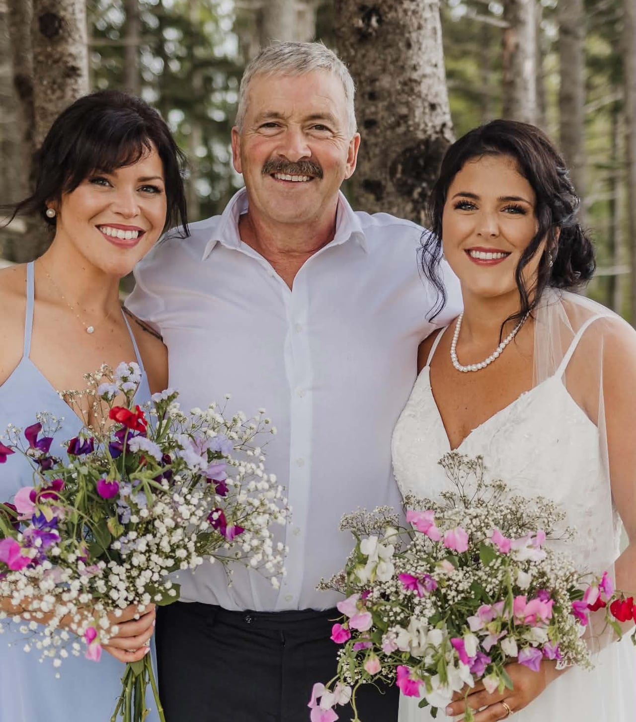 A man smiles between two women dressed as brides and bridesmaids.