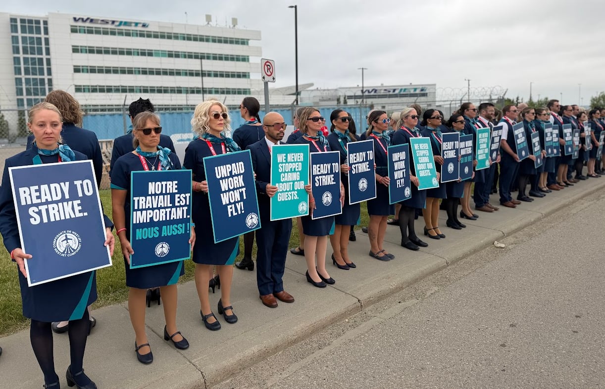 Flight attendants in blue and teal uniforms hold up signs that say "Ready to Strike" and "Unpaid Work Won't Fly" in front of WestJet's head office in Calgary.