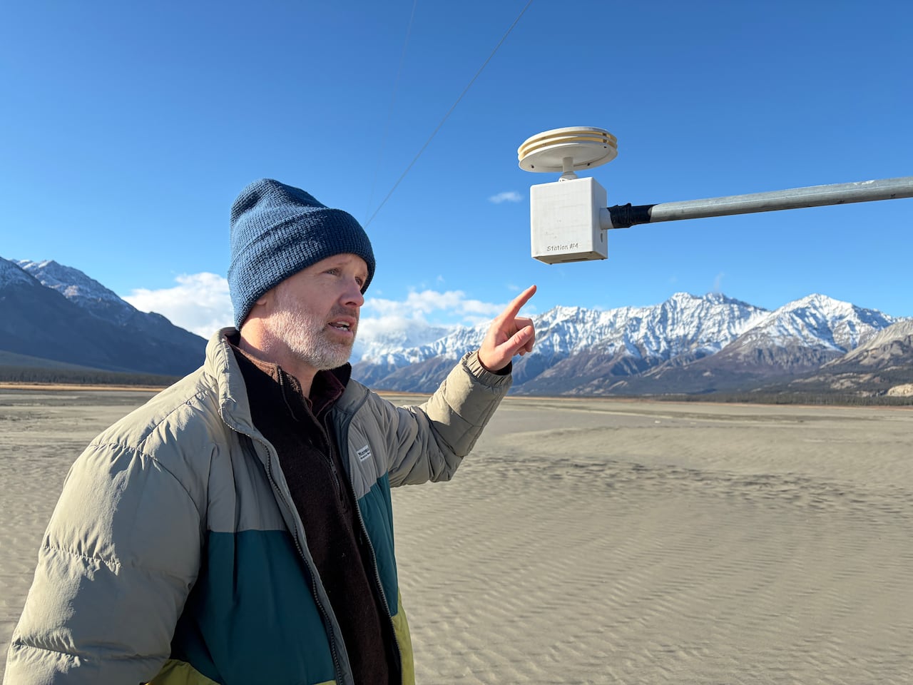 James King points to a particle sensor mounted on a metal arm above the exposed Ä'äy Chù (Slims River) Valley floor, with snowy mountains behind him.