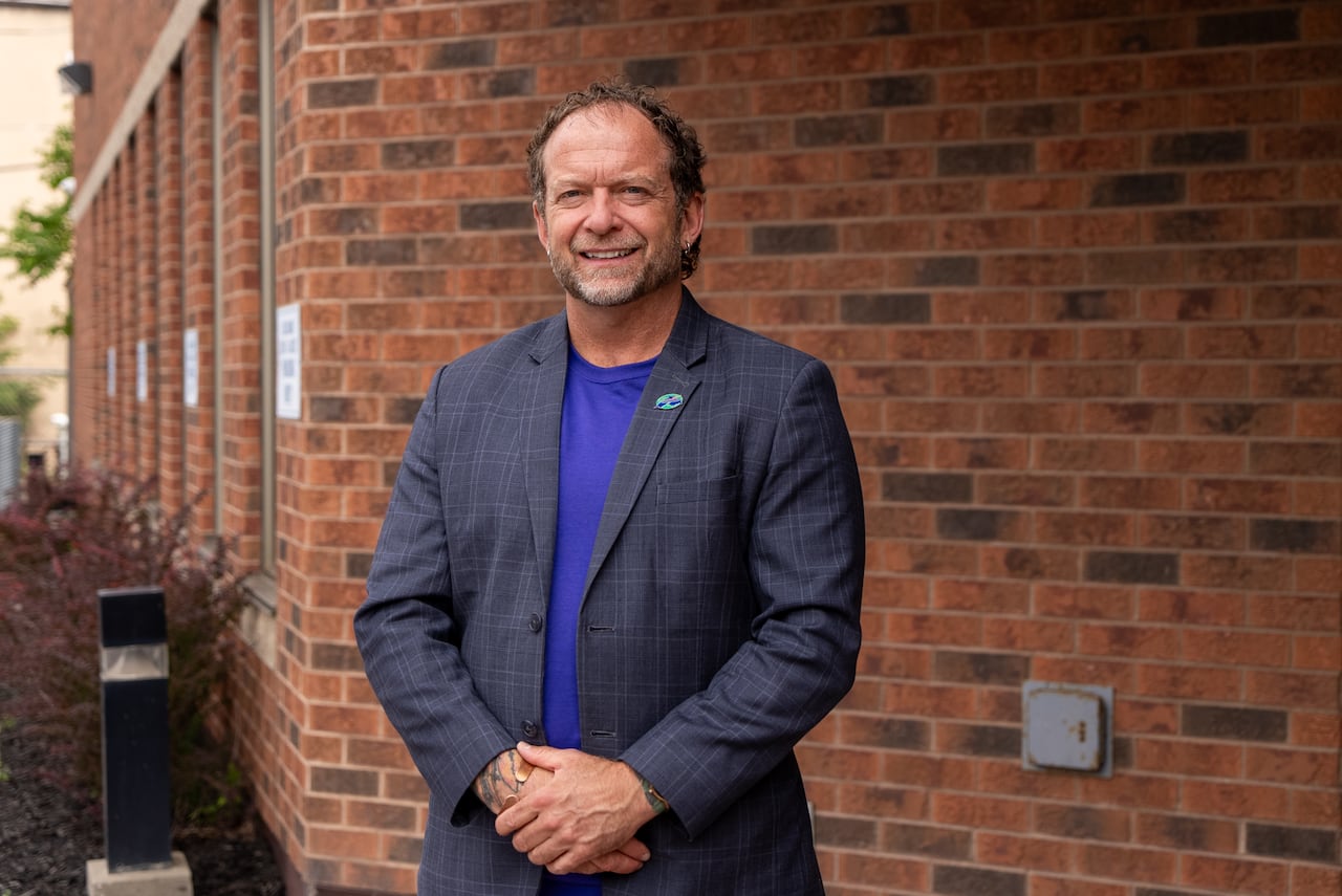 A person wearing a blazer stands in front of a brick building.