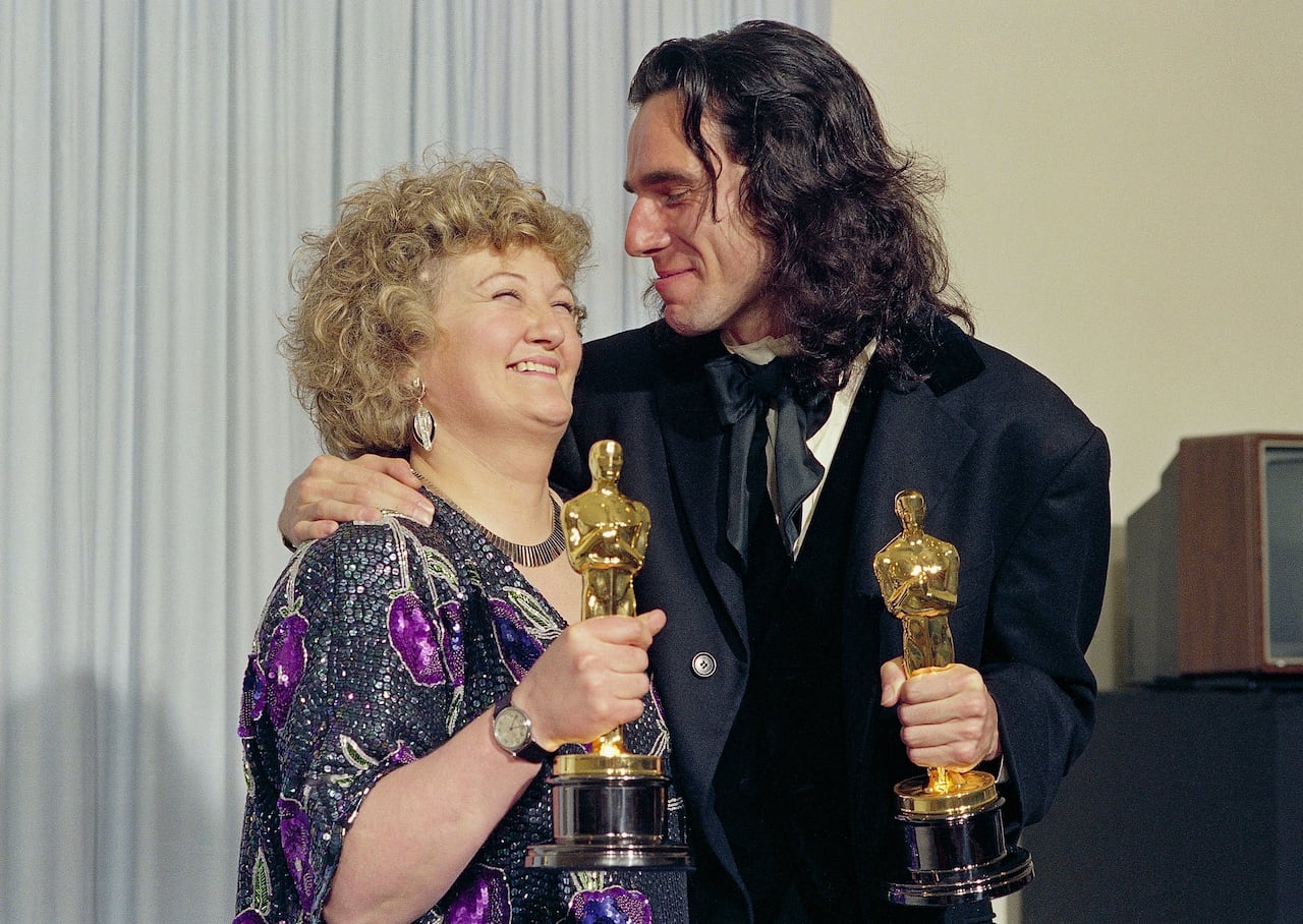 A smiling woman waring a sequined dress holds a golden Oscar statuette, as a smiling man also holding an Oscar trophy wraps an arm around her.