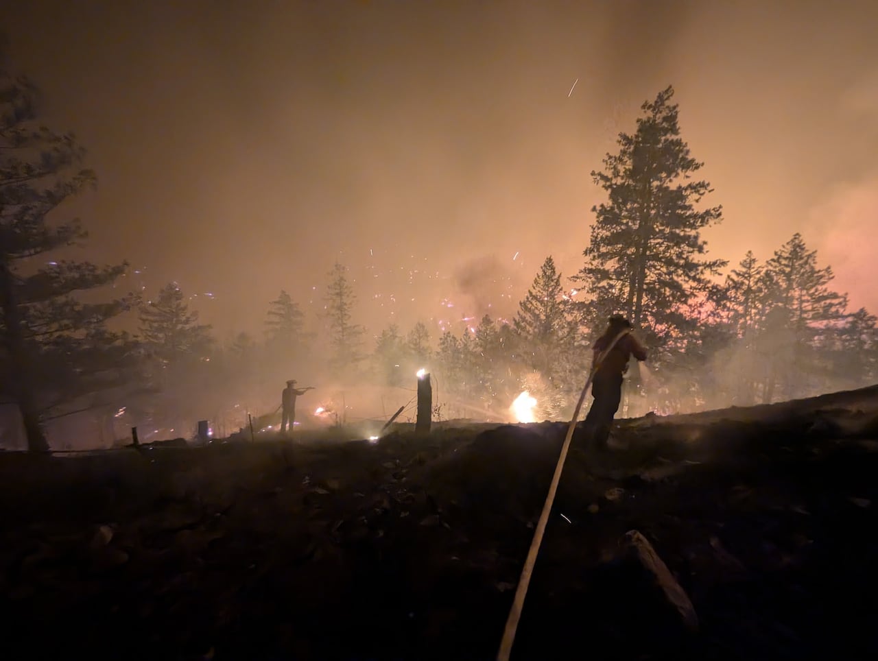 A firefighter pulls a hose through a smoky landscape at night.