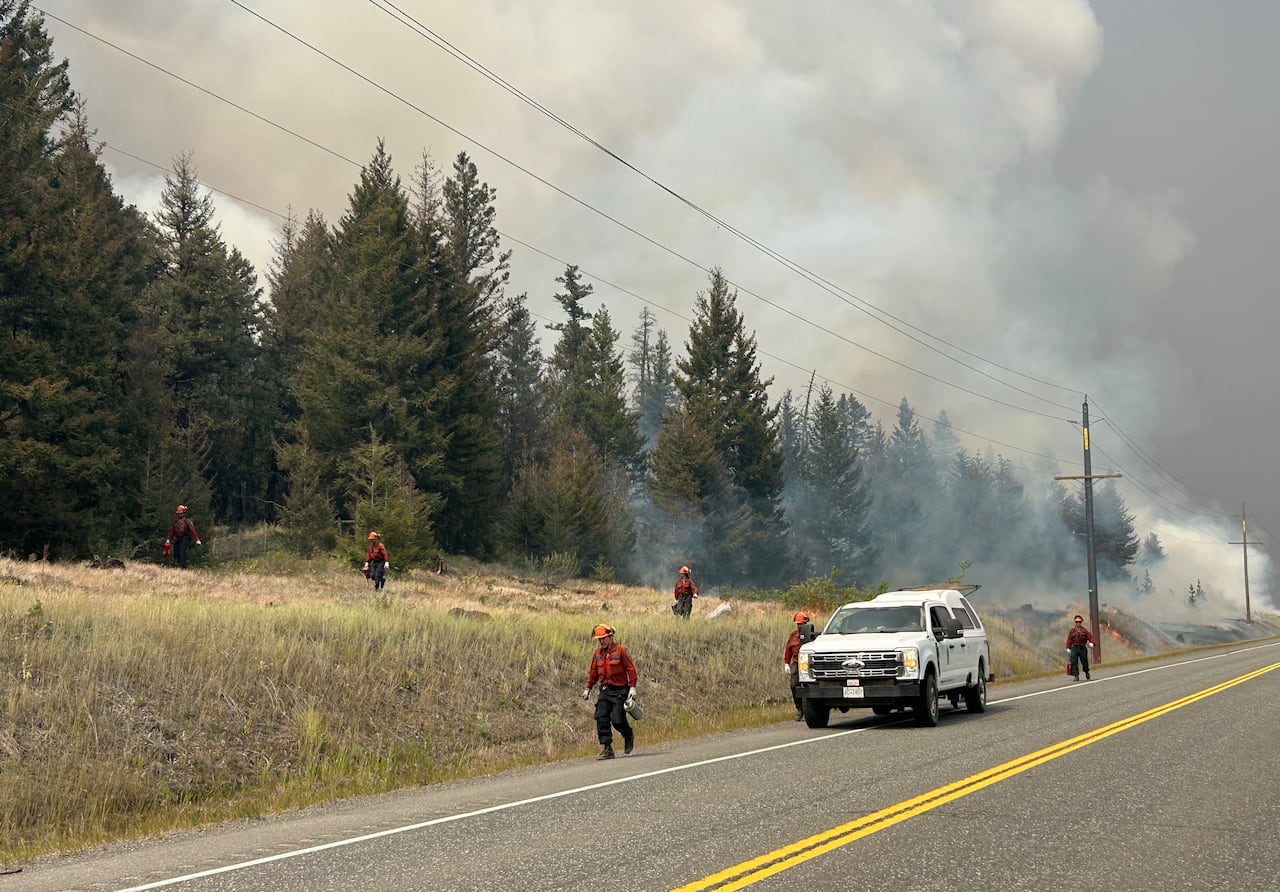 Six people in red uniforms carrying hand torches walk on a grassy area beside a highway amid smoke and flames. There is a white truck parked on the road.