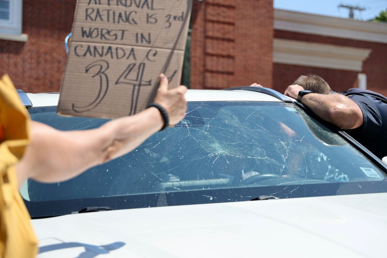 person holds cardboard sign in front of a smashed windshield
