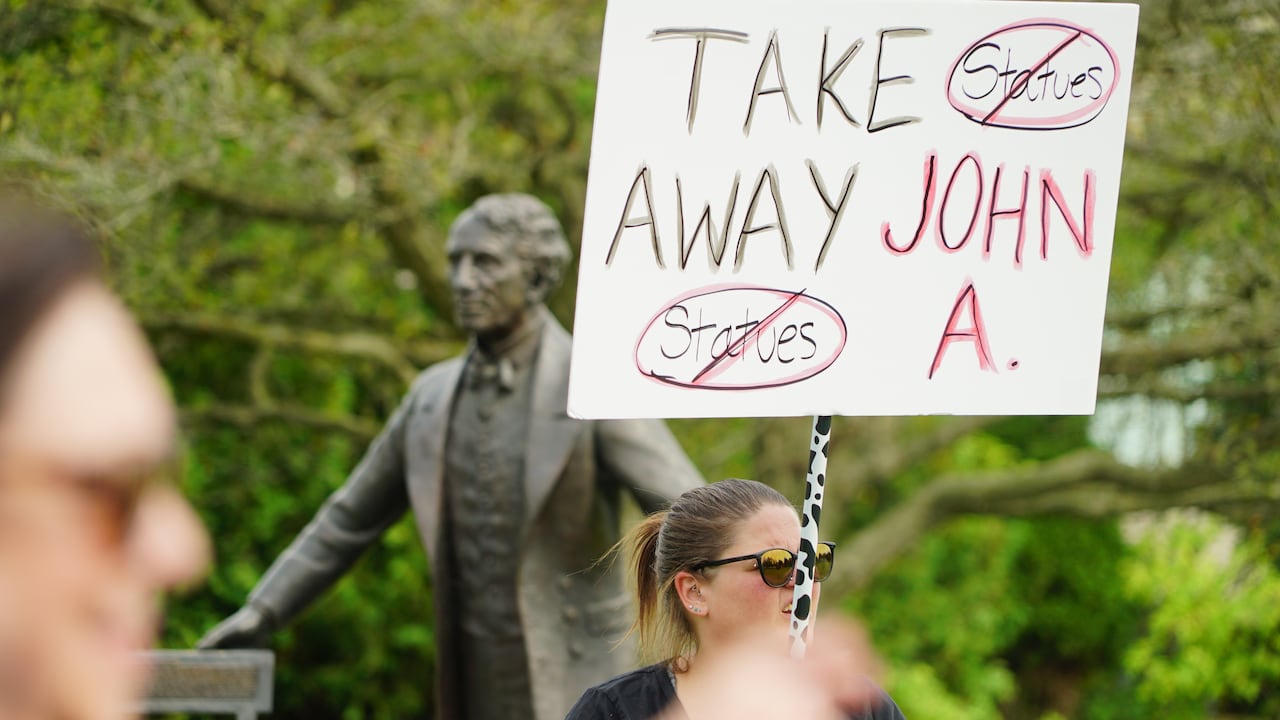 A protester holds up a sign that says, 'take away John. A.' Behind the protester is Sir John A. Macdonald's statue.