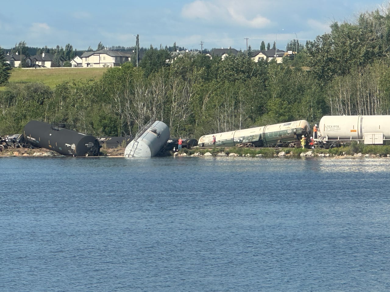 Multiple emergency responders in neon jackets wander around the scene of a derailed train that is partially submerged in water.