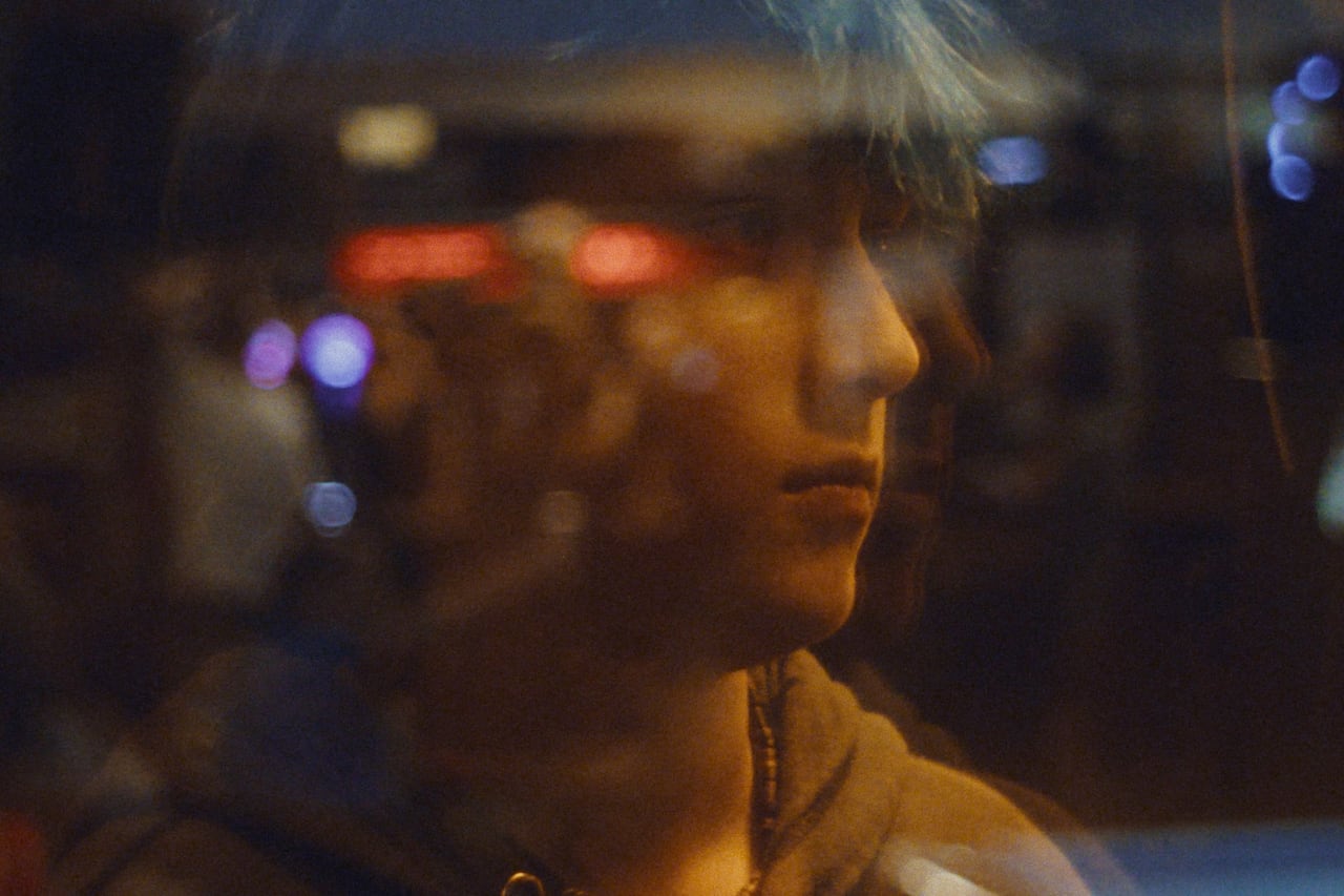 A boy with blue hair looks off into the distance behind a window pane.