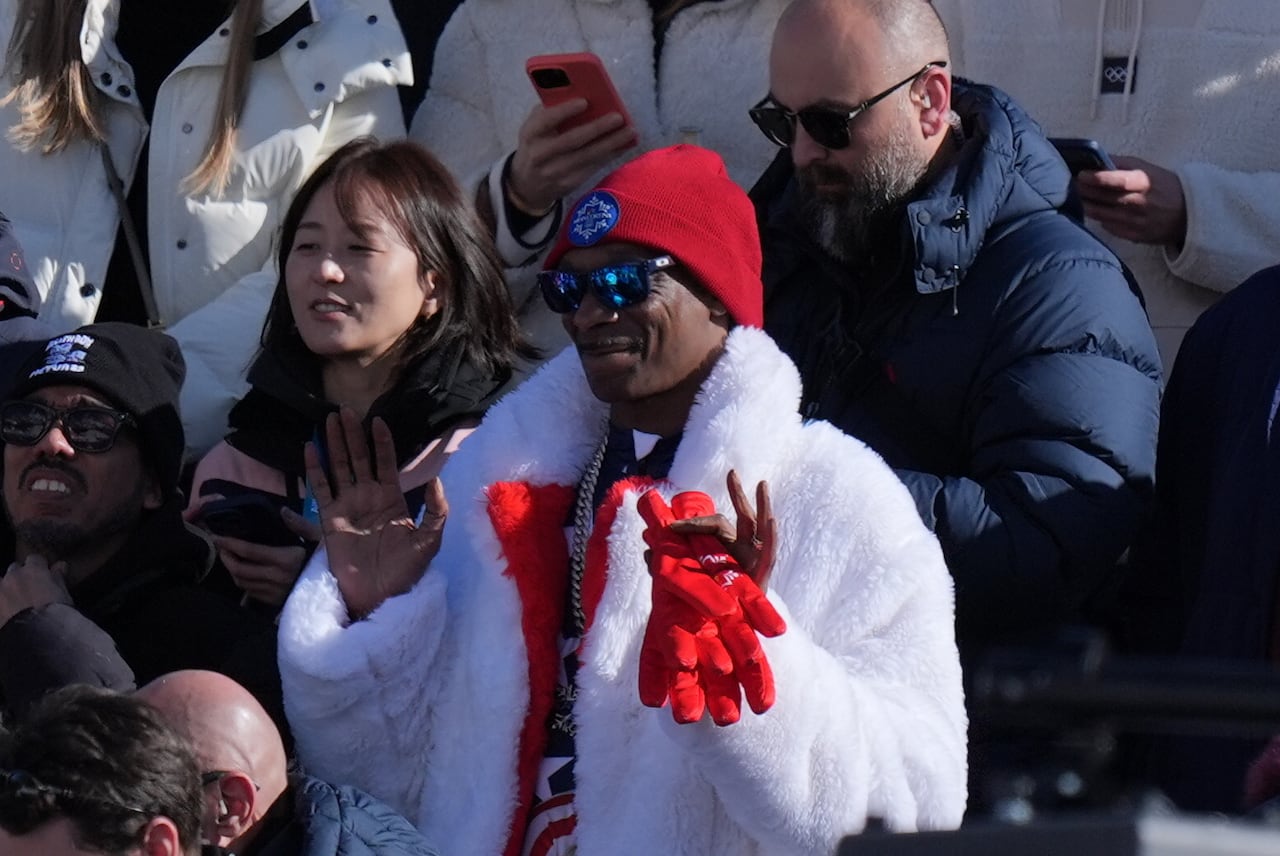 A man standing in a crowd in a white fur coat holding red gloves.