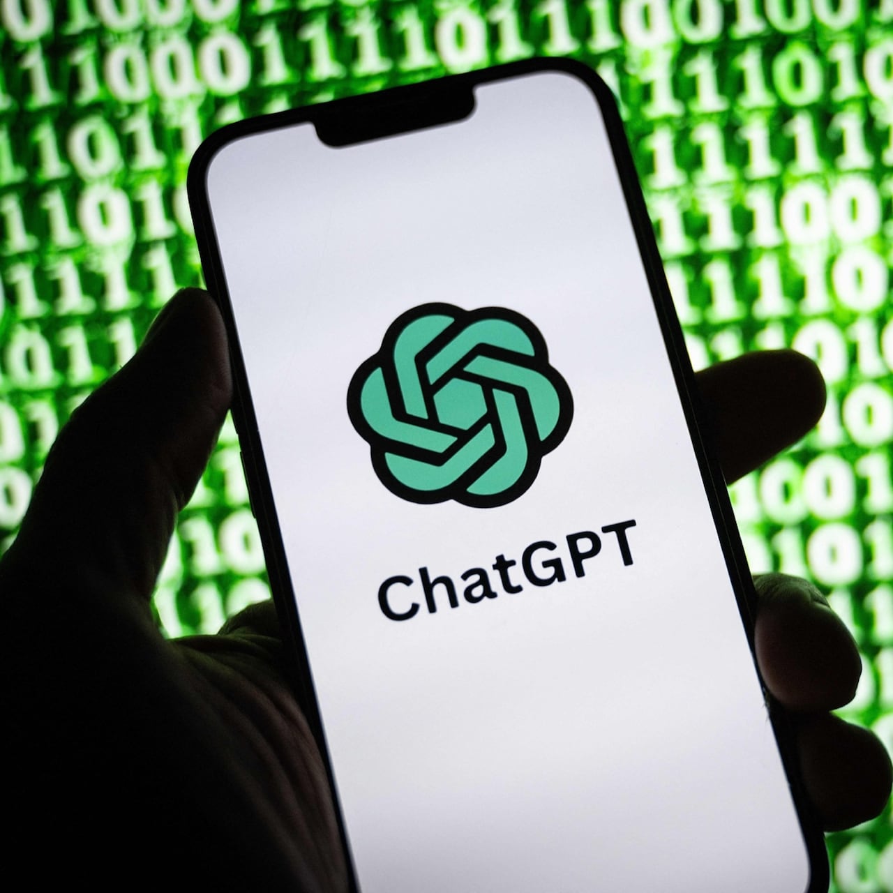 A hand holds up a phone displaying the ChatGPT logo in front of a wall of binary code.