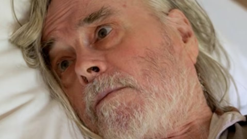 A close up of an older man with long hair and a beard in a bed.