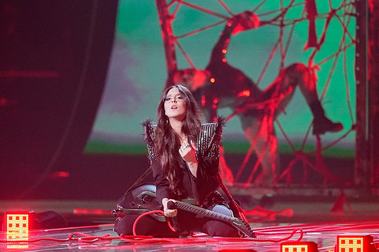 A woman holding a guitar is on her knees on stage. In the background, a man appears to be caught in a spiderweb.