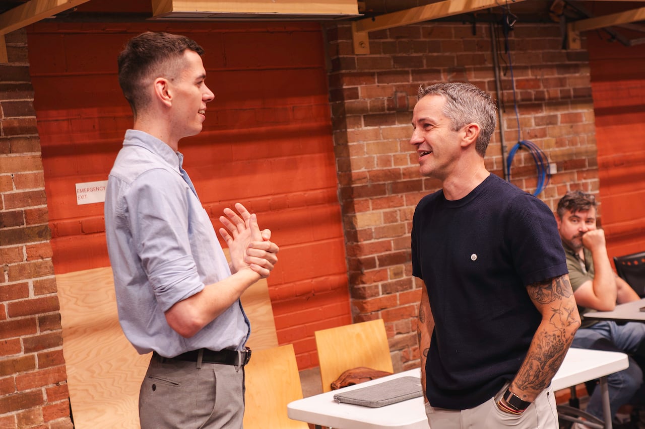 A tall man in a button-down shirt speaks to a shorter man wearing a t-shirt that reveals tattoos. They are in a room with brick walls.