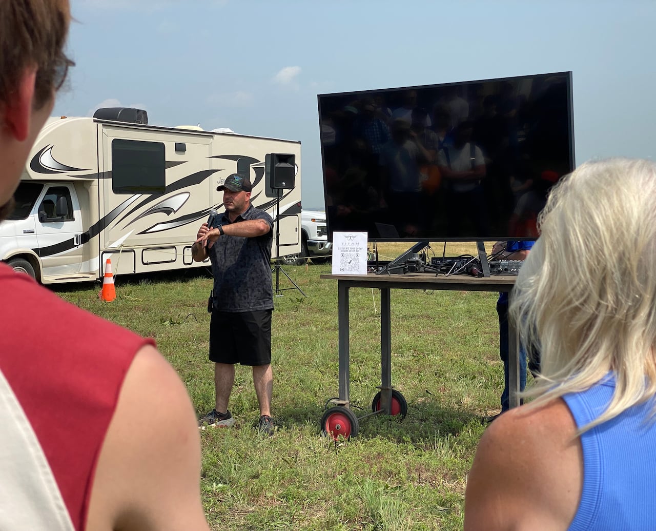 The backs of two people are in the foreground. In the middle of them, a man stands further ahead speaking into a microphone in front of a TV screen in a field.