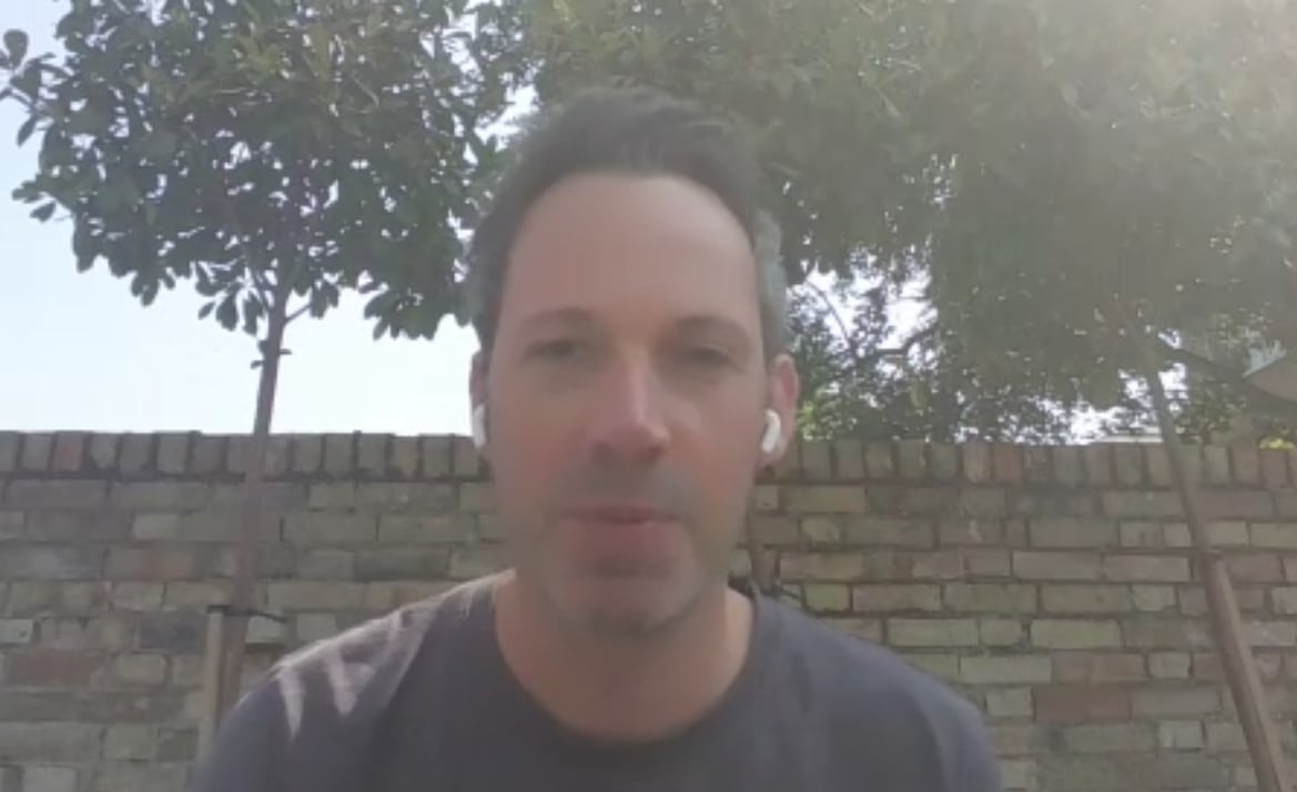 A Zoom screenshot of a man sitting outside, wearing wireless earbuds.
