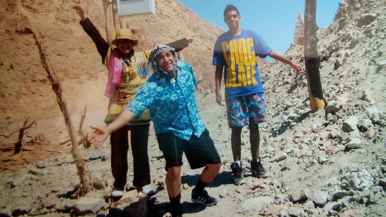 A photo of a family posing on a rocky hill, with a teen boy on the left wearing summer clothes next to his dad, and mom, both wearing colorful summer attire.