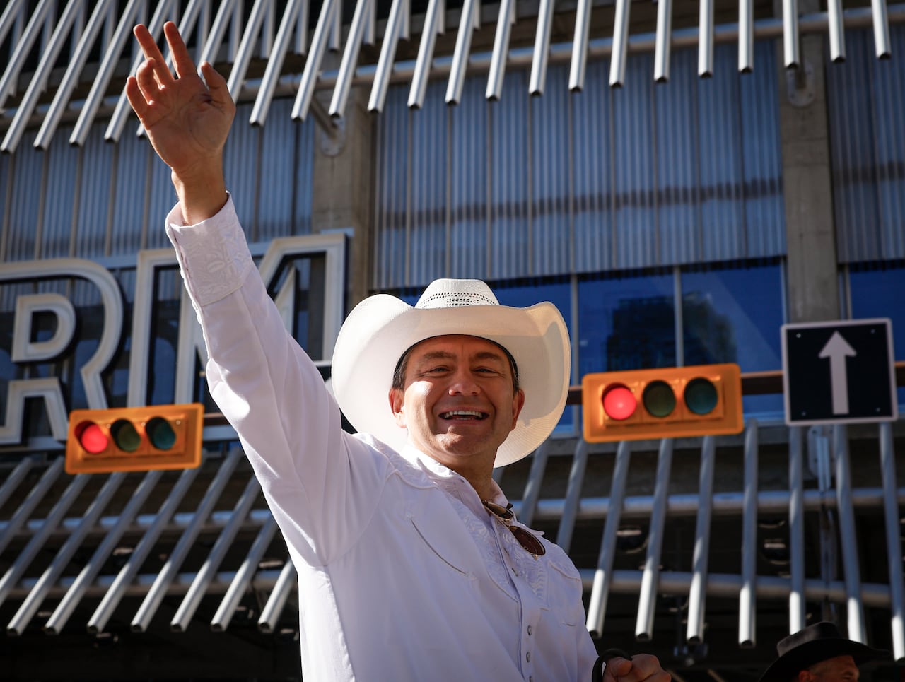 Pierre Poilievre is seen wearing a white cowboy hat and waving to a crowd during the Calgary Stampede parade. The horse he is riding on is out of frame.