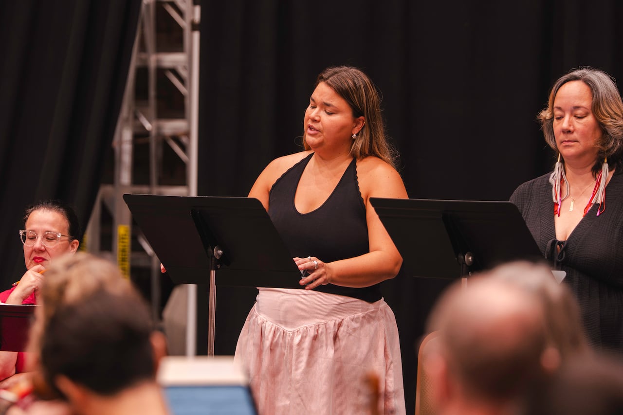 A woman in a pink dress and black halter top sings with a music stand before her. There’s another woman standing beside her behind another music stand.