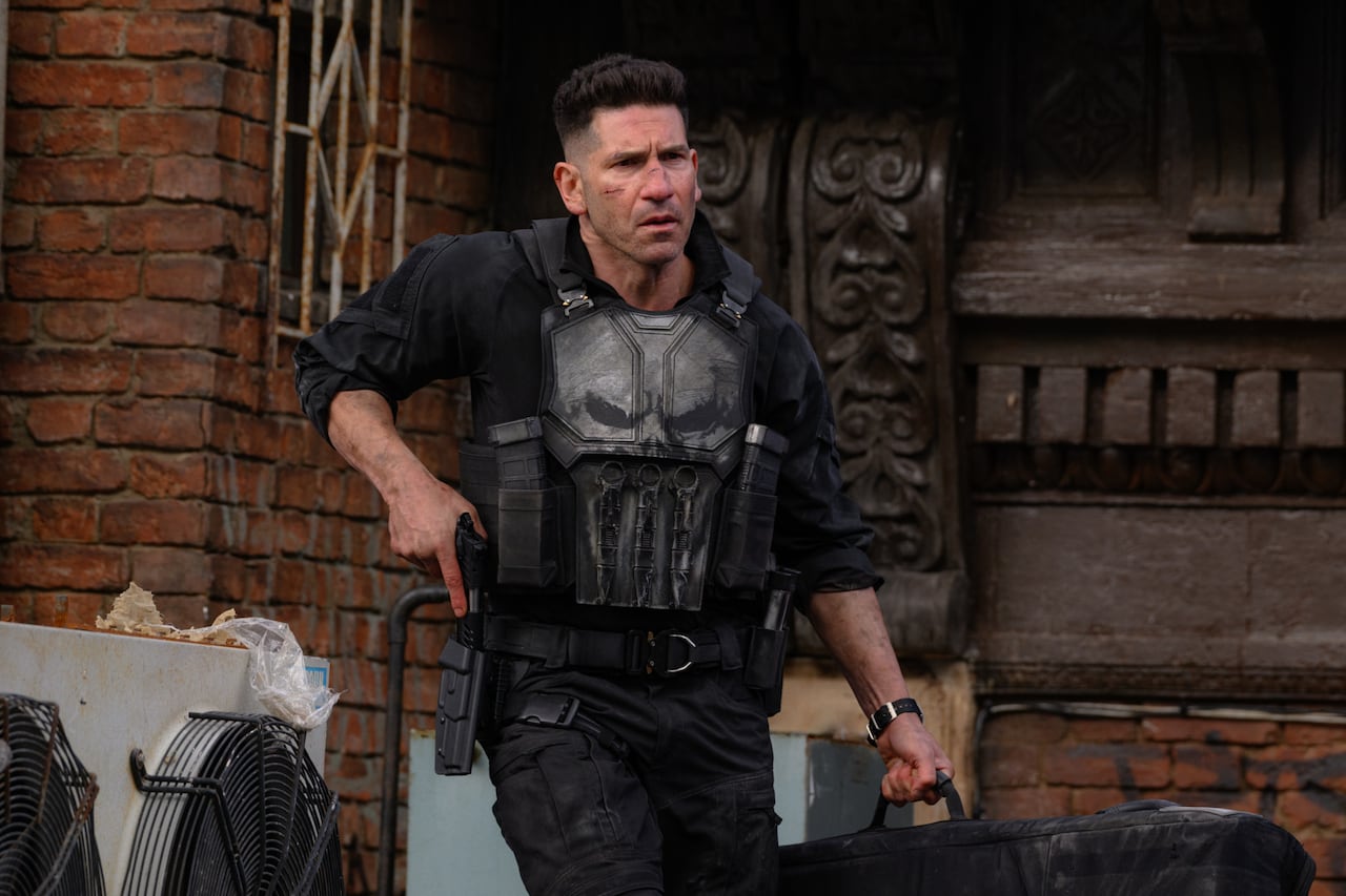 Actor Jon Bernthal is dressed as The Punisher, wearing combat gear with a skull insignia as he grimaces and pulls a pistol out of a holster.