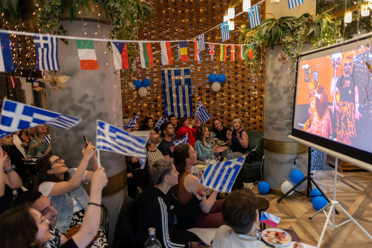 Flags from countries around the world hang from the roof of a room, while a group of people waving blue and white Greek flags sit in front of a TV.