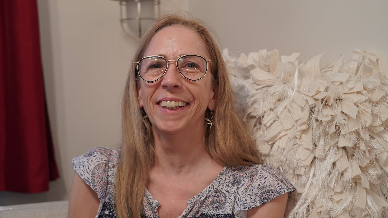 A woman with blonde hair and glasses smiles at the camera from her seat on a couch.