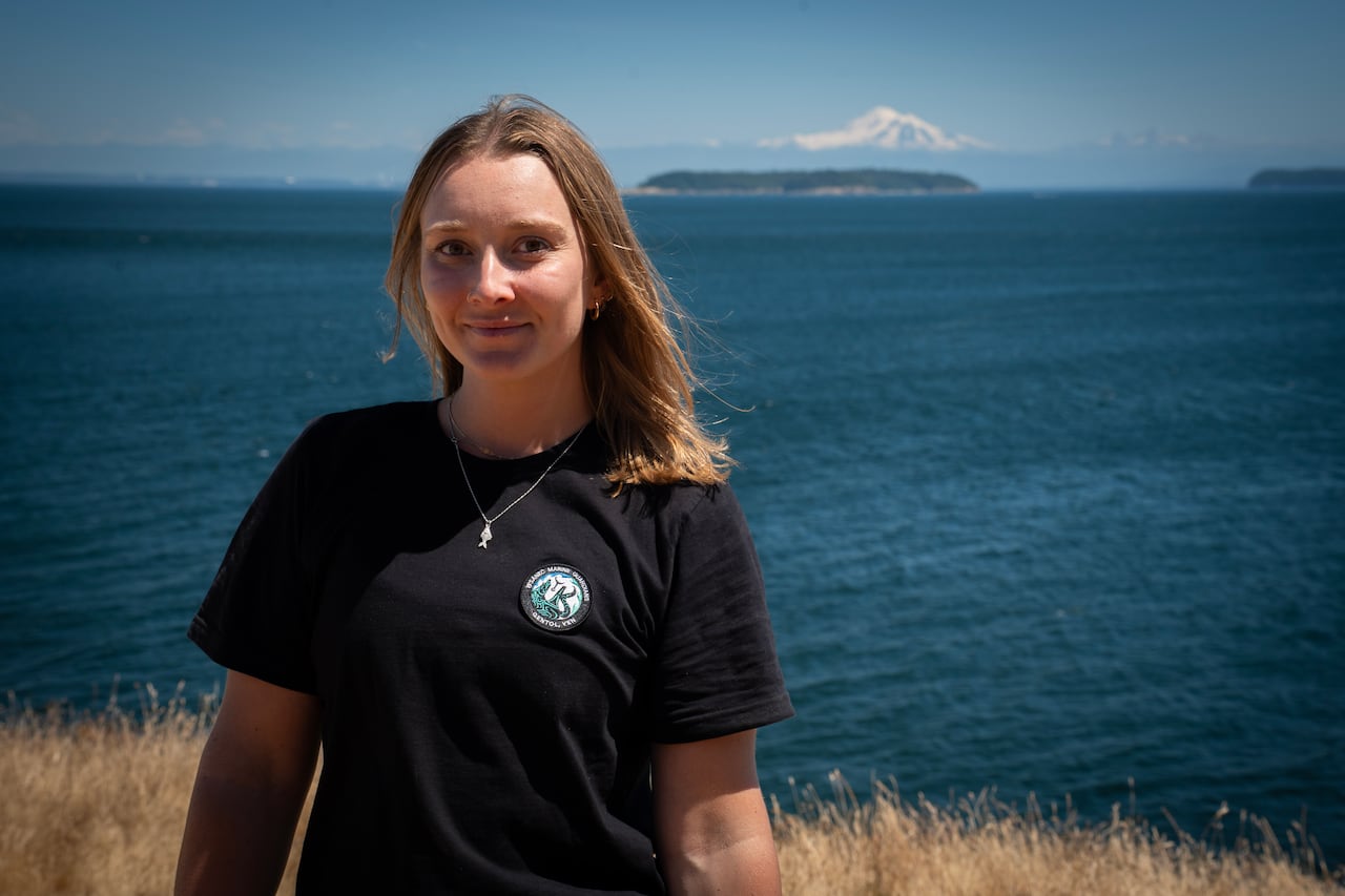 Rachel is a 20-something year old white woman with blonde shoulder length hair. She is standing on the shore of an island with the Salish Sea in the background. The sky is blue. She is smiling.