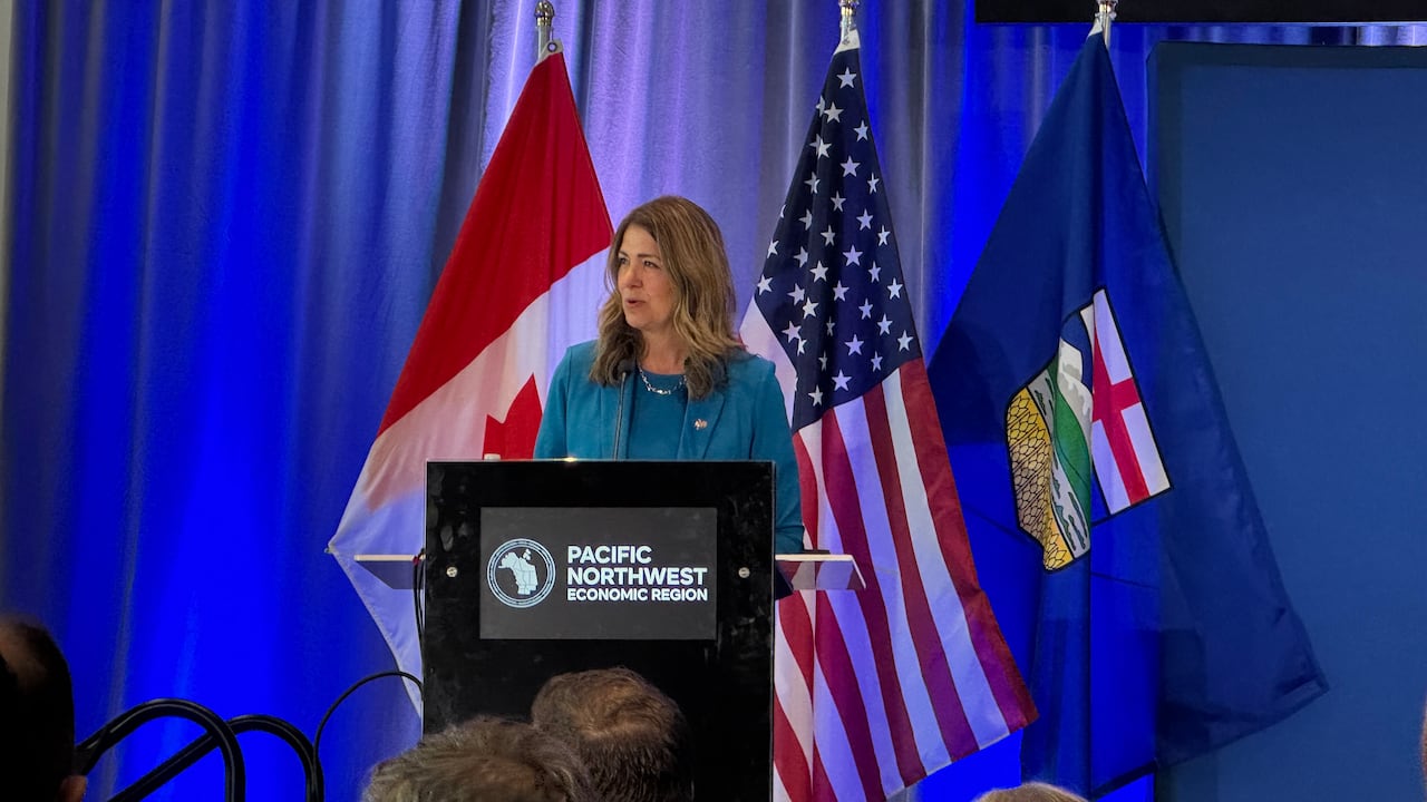 A woman with brown hair speaks into a microphone at a podium. Behind her are flags for Canada, the U.S., and Alberta. The podium has a logo on it that reads "PACIFIC NORTHWEST ECONOMIC REGION." She is wearing a blue blazer with a matching dress.