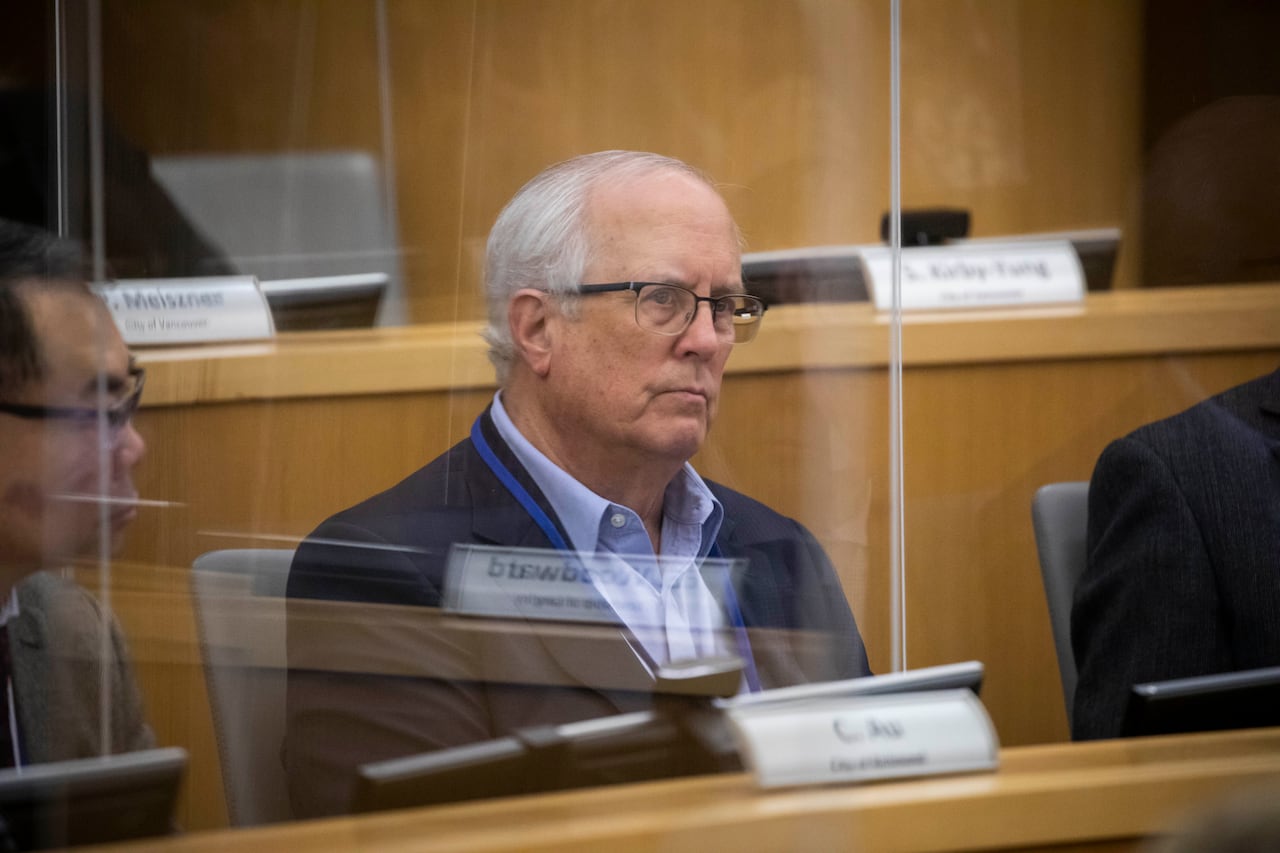 A white man sits in a boardroom and looks on, next to an East Asian man.