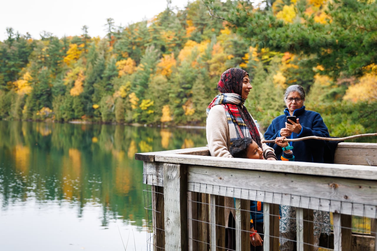 Two women standing on a platform that overlooks a body of water rimmed by forest displaying fall foliage.