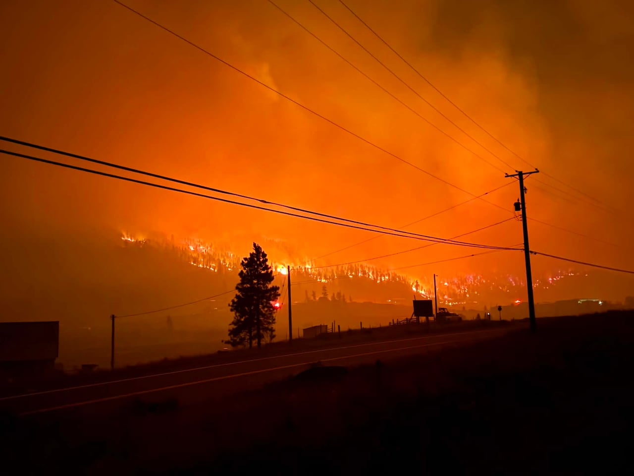 Fire blazes in a forest across from a highway at night.