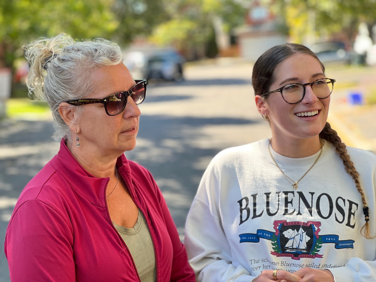 Two individuals stand on a street. One wears a pink sweater with sunglasses. The other wears a white sweater.