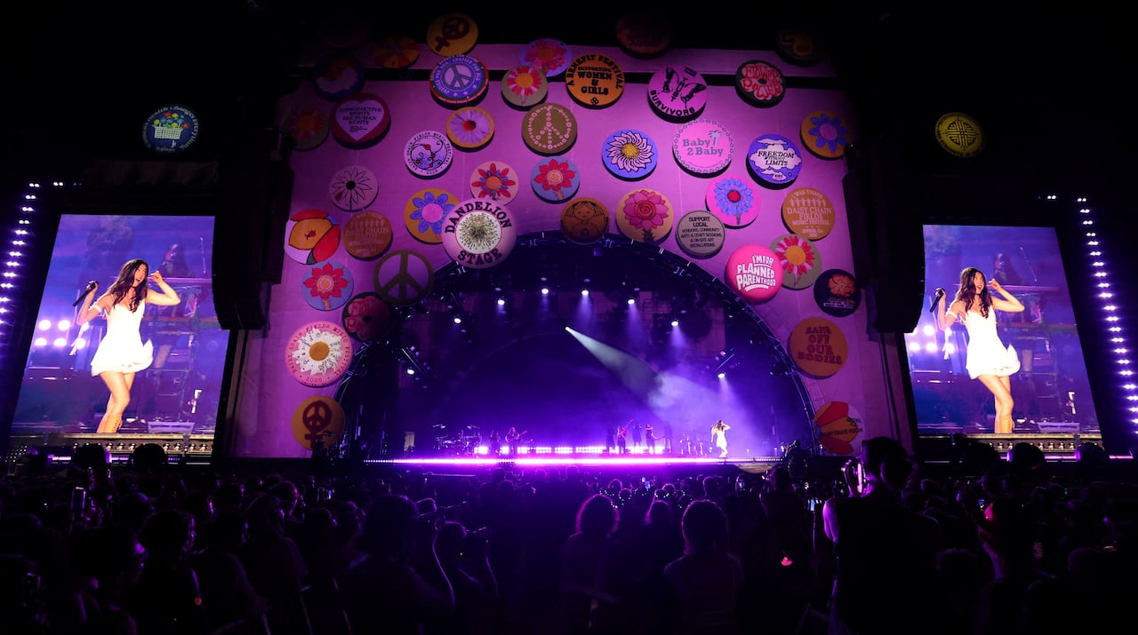 A large stage is shown at night, with two screens on either side showing a woman in a white dress.